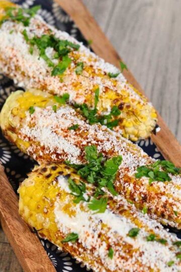 three elotes (Mexican street corn) on wooden blue plate with lime on the side
