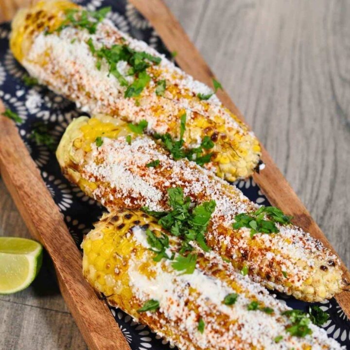 three elotes (Mexican street corn) on wooden blue plate with lime on the side