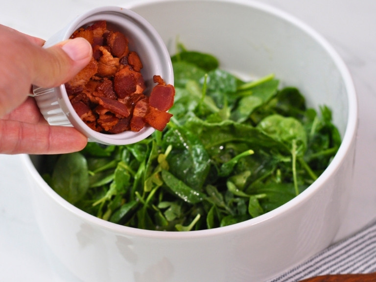 Arugula and Spinach in white bowl with bacon being mixed in.