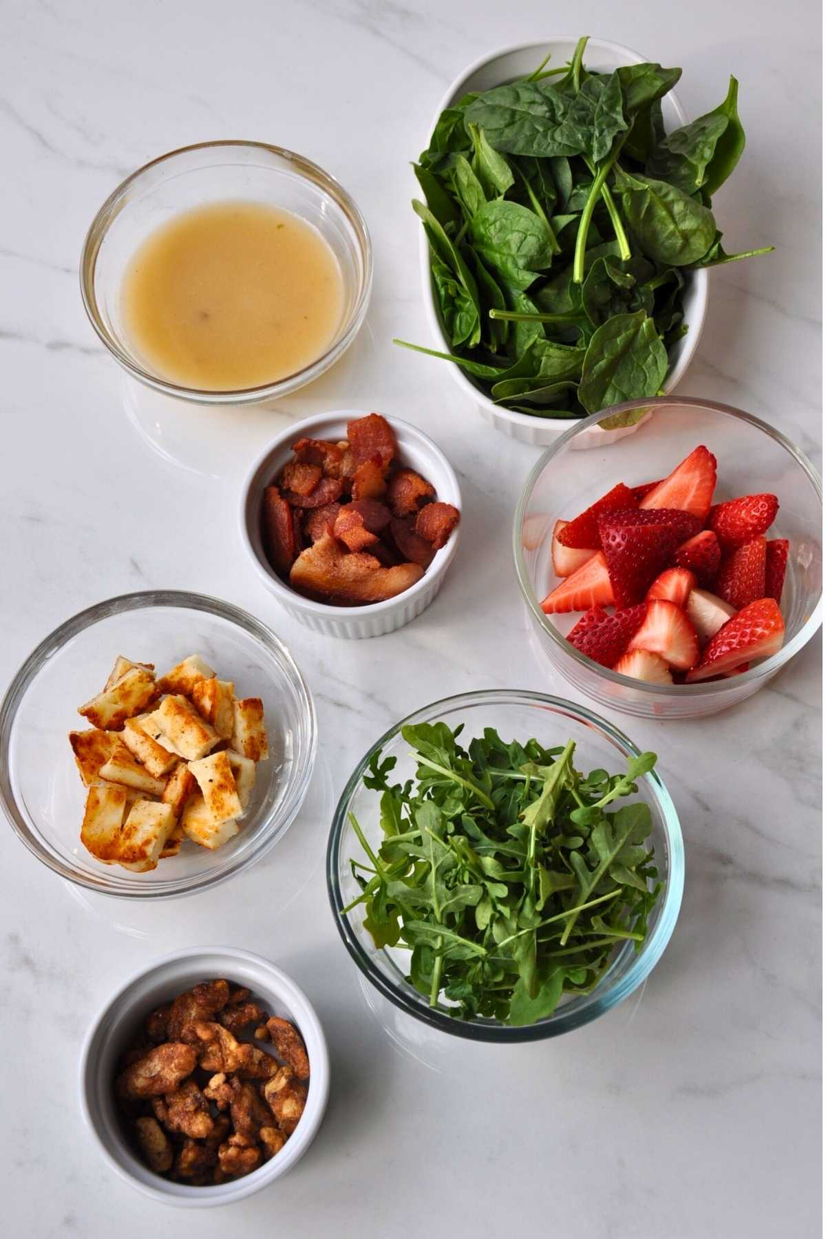 small glass bowl of vinaigrette, Halloumi grilled cheese, cut strawberries, bowl of chopped cooked bacon and candied walnuts, white bowl spinach and glass bowl of arugula