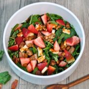arugula, spinach, strawberries, Halloumi grilled cheese and bacon in white salad bowl with brown wooden spoon on wooden on wood table