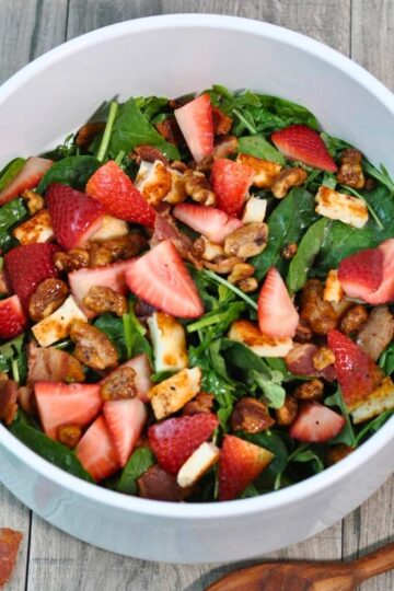 arugula, spinach, strawberries, Halloumi grilled cheese and bacon in white salad bowl with brown wooden spoon on wooden on wood table