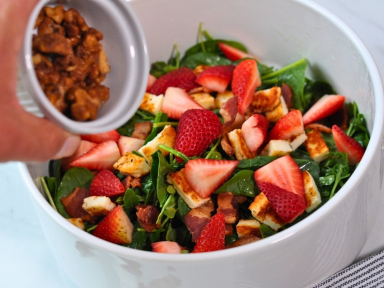 Arugula and Spinach Salad, strawberries and Halloumi grilled cheese in white salad bowl with brown wooden spoon. candied walnuts being poured into salad.
