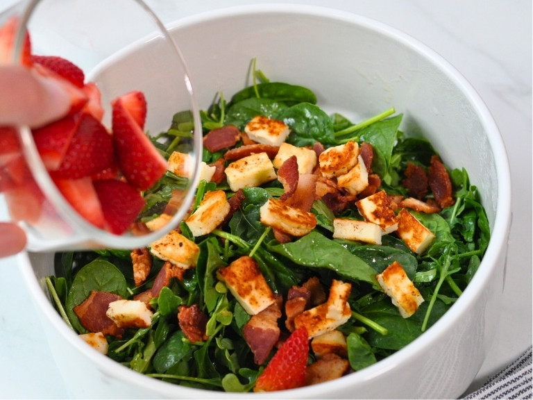 Arugula and Spinach Salad, strawberries and Halloumi grilled cheese in white salad bowl with brown wooden spoon. strawberries being poured into salad.