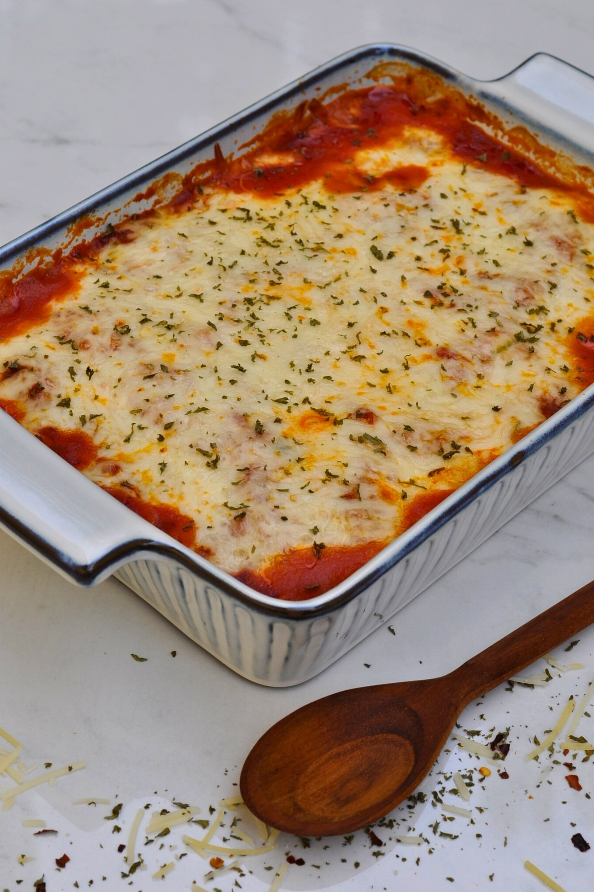 beef and spinach lasagna in white and blue baking dish on marble countertop with wooden spoon