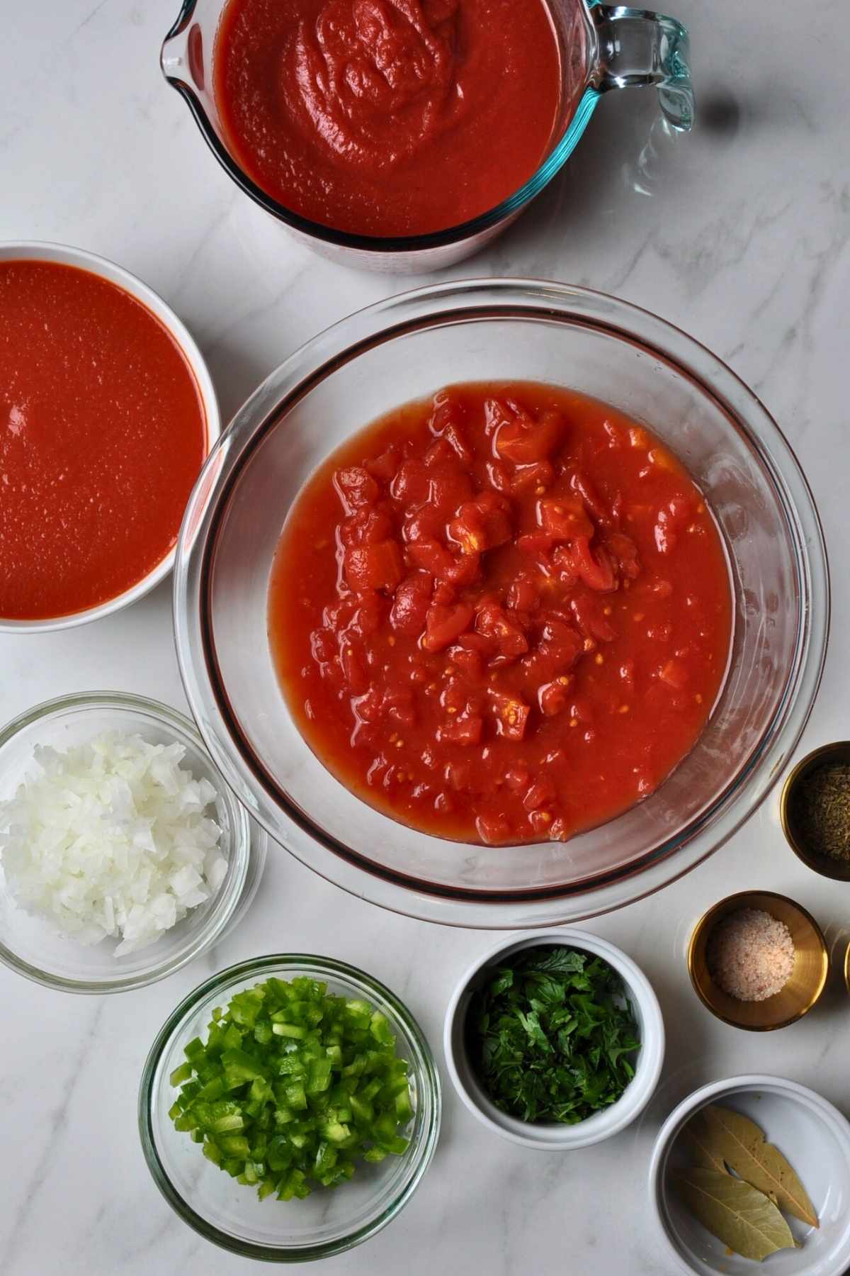 diced tomatoes in glass bowl, tomato sauce in bowl, crushed tomatoes in glass bowl, white onion in glass bowl, diced green bell pepper in glass bowl, chopped Italian parsley in white ramekin, 2 bay Leaves in white ramekin, seasoning salt in gold ramekin.