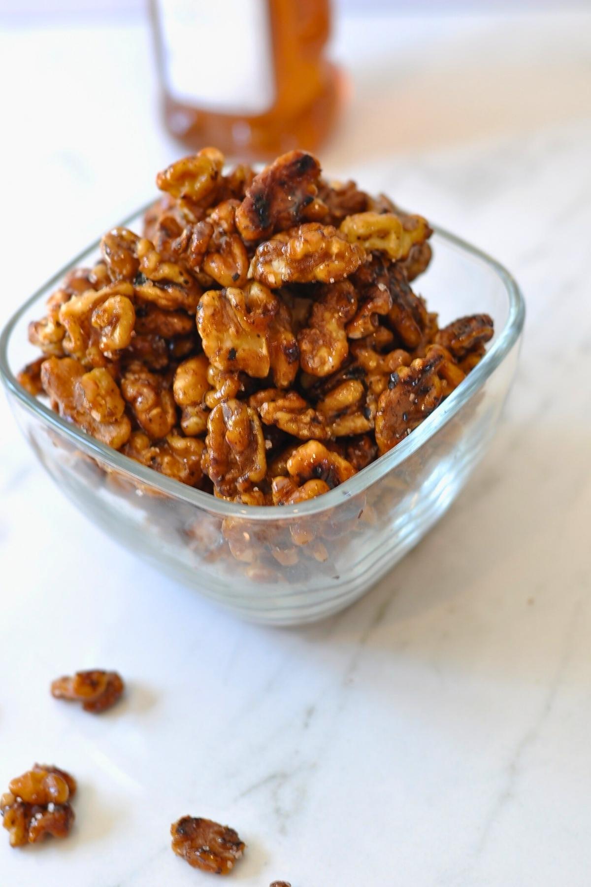 glazed walnuts in a glass bowl. three walnuts are sitting on the counter with a bottle of honey in the background