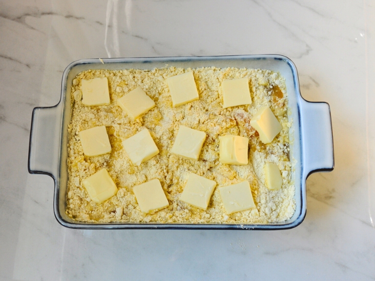 un cooked peach cobbler in blue and white baking dish with 14 butter squares on top