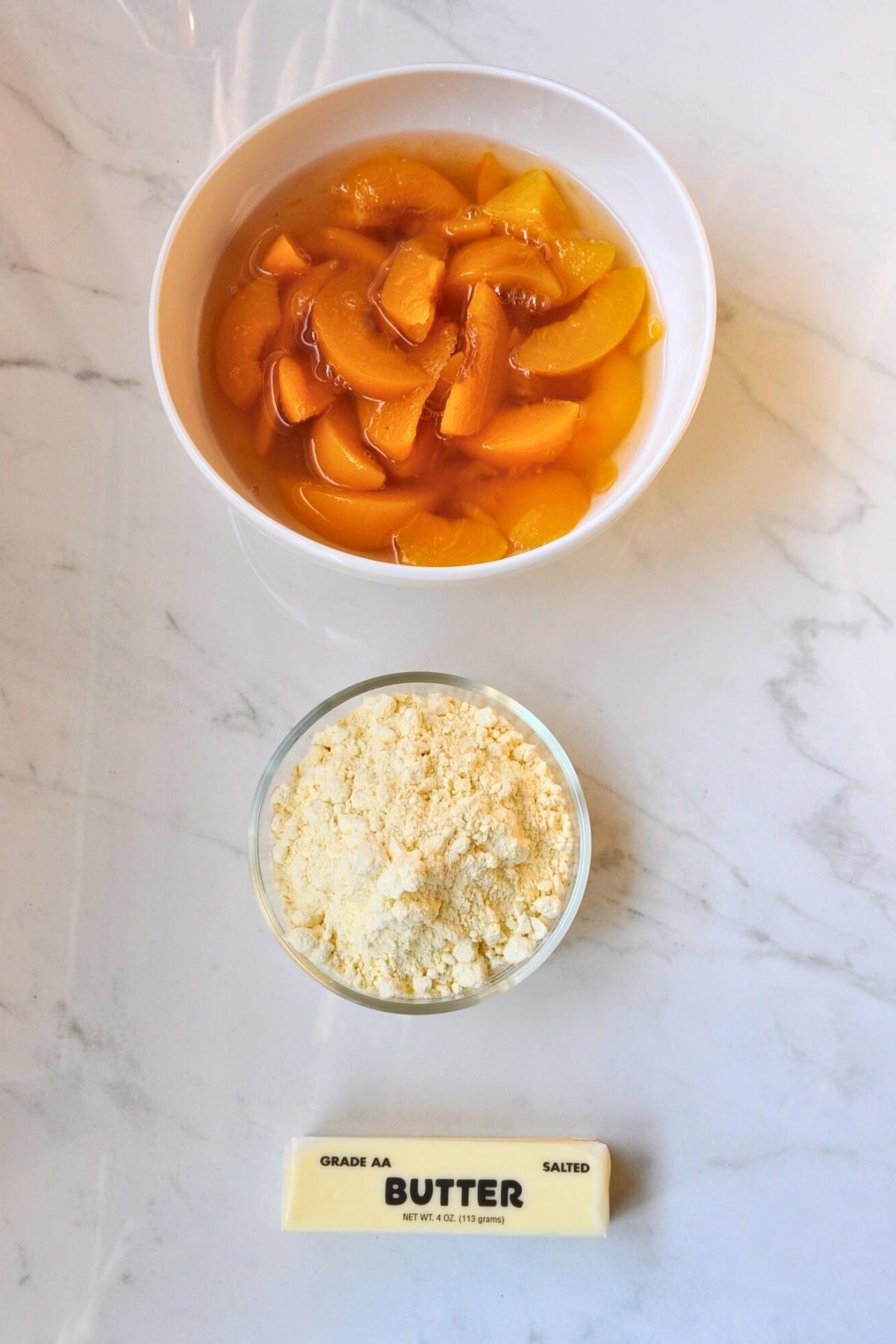 white bowl of sliced canned peaches, glass bowl of yellow cake mix and a stick of butter in wrapper on countertop