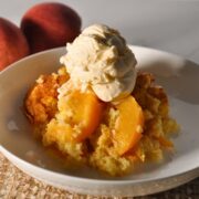 peach cobbler with scoop of vanilla ice cream on a white plate with two whole peaches in the background