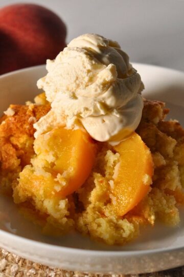 peach cobbler with scoop of vanilla ice cream on a white plate with two whole peaches in the background